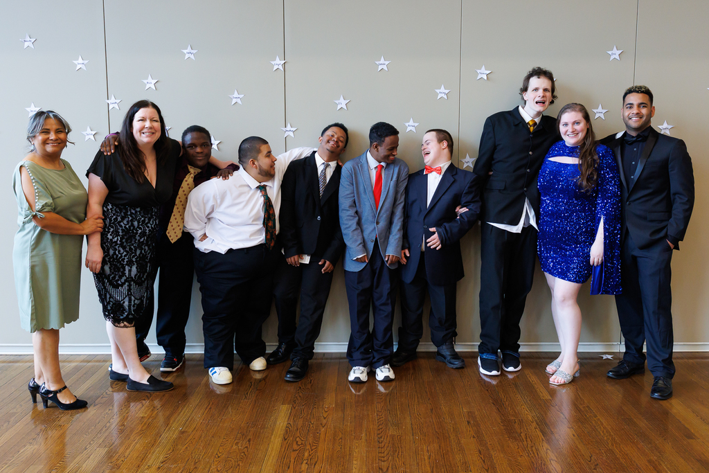 A class of ten people, students and staff, stand together for a photo while smiling and laughing alongside each other.