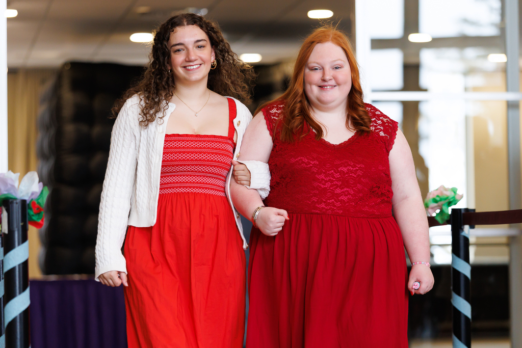 Two young women in red dresses walk arm-in-arm, smiling.