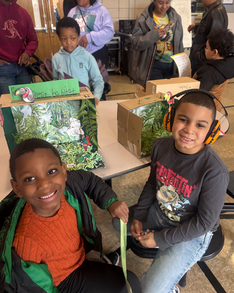Two student look at the camera for a photo while sitting near their 3D Habitat.