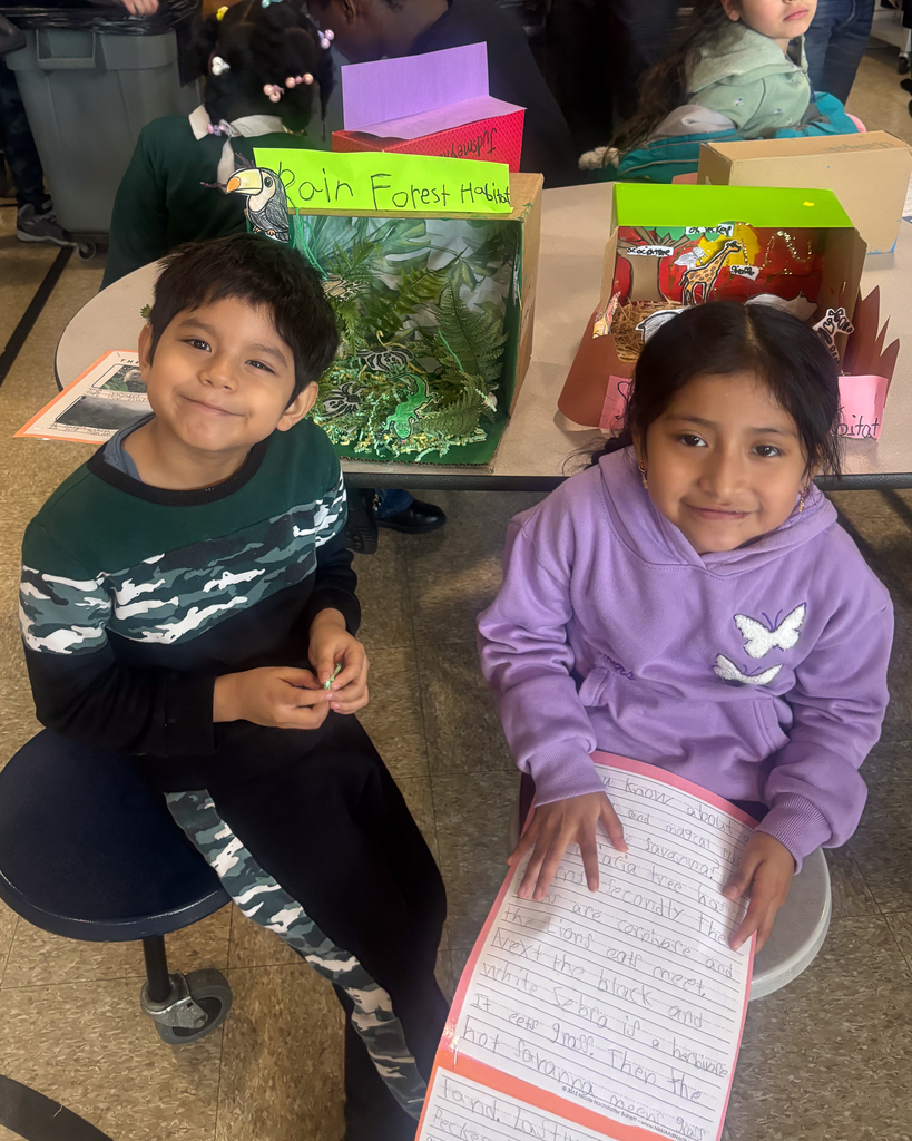 Two student look at the camera for a photo while sitting near their 3D Habitat.