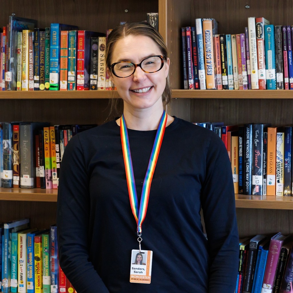 A woman with glasses smiles in front of a bookshelf filled with books.