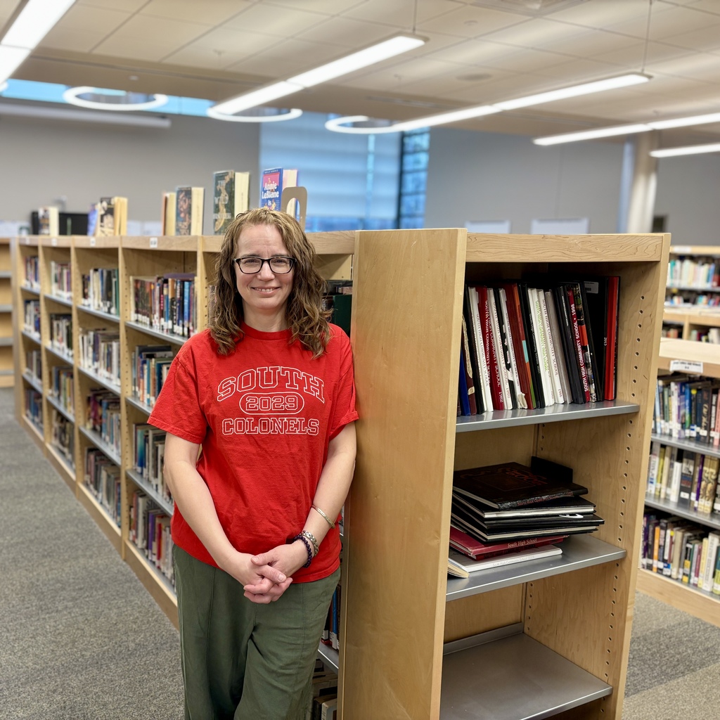 A woman wearing glasses and a red t-shirt with 'SOUTH 2029 COLONELS' stands in a library aisle.