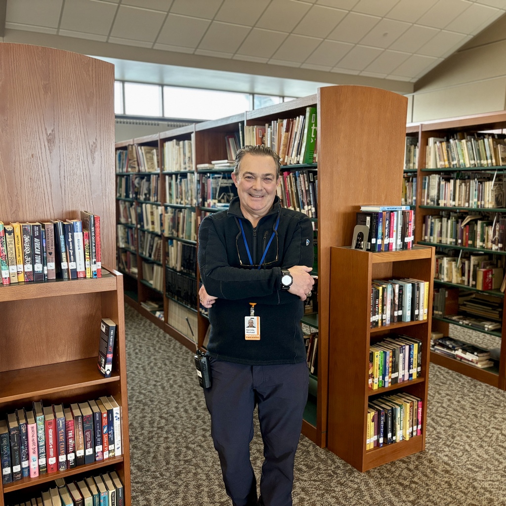 A man with a name tag reading 'Garvey, Michael' stands with his arms crossed in a library aisle, surrounded by bookshelves.