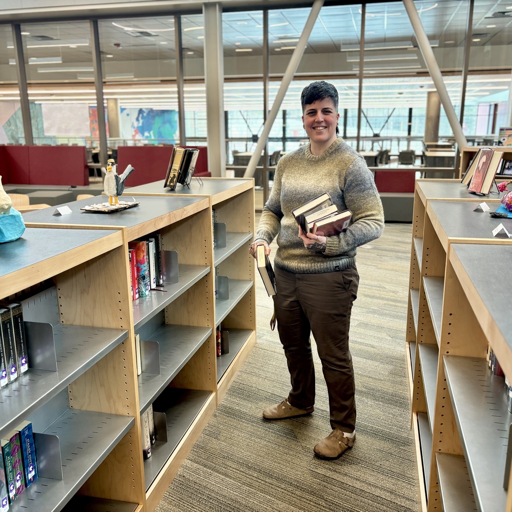 A person smiles while holding several books in a library aisle with bookshelves.