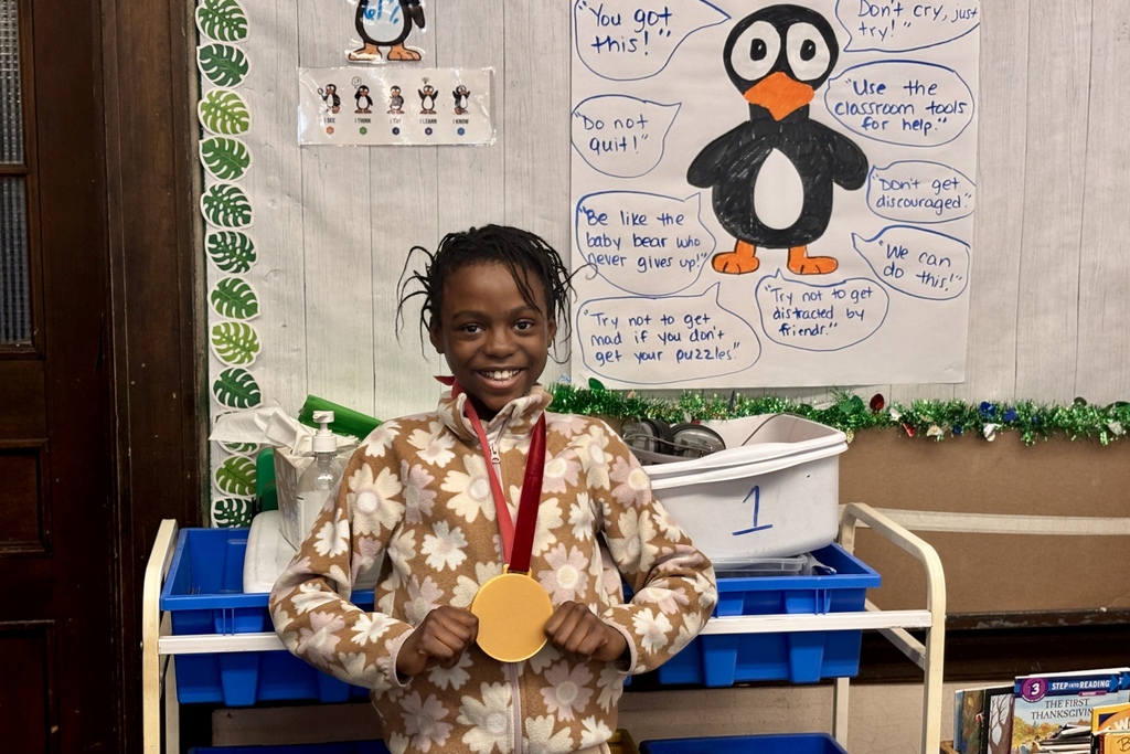 A smiling young person proudly holds a gold medal in a classroom decorated with penguin-themed motivational posters.