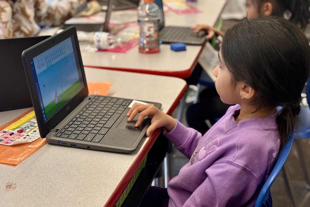 A young student in a purple sweatshirt uses a laptop in a classroom setting.