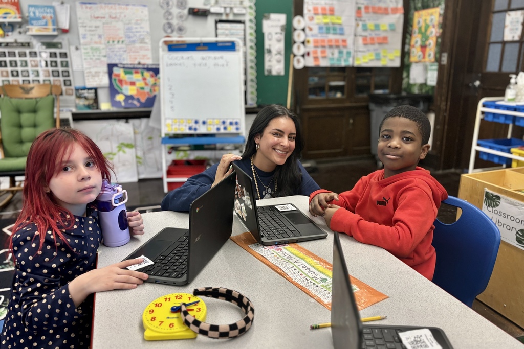A teacher smiles at the camera while two students work on laptops in a classroom.