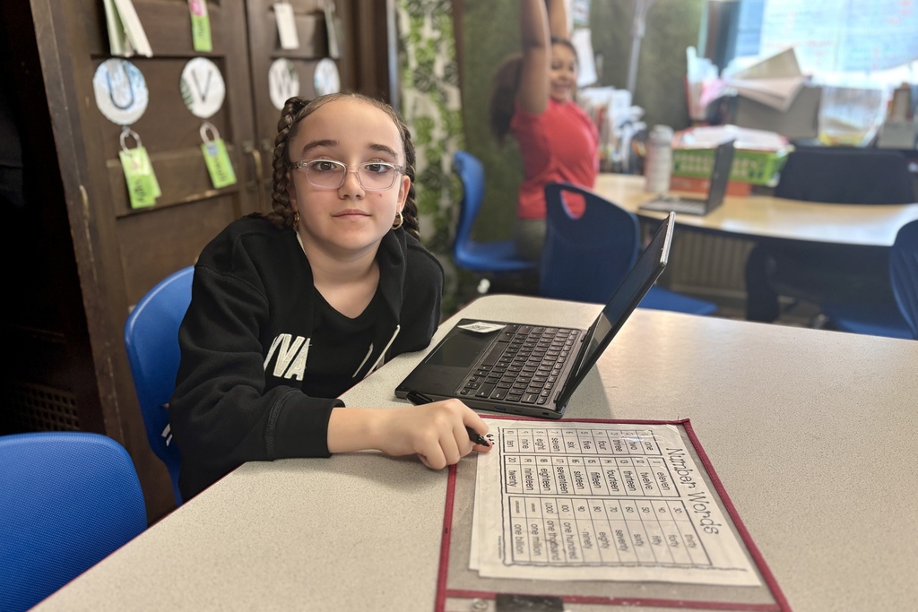 A young student with glasses and braided hair looks at the camera while sitting at a desk with a laptop and a number words chart.