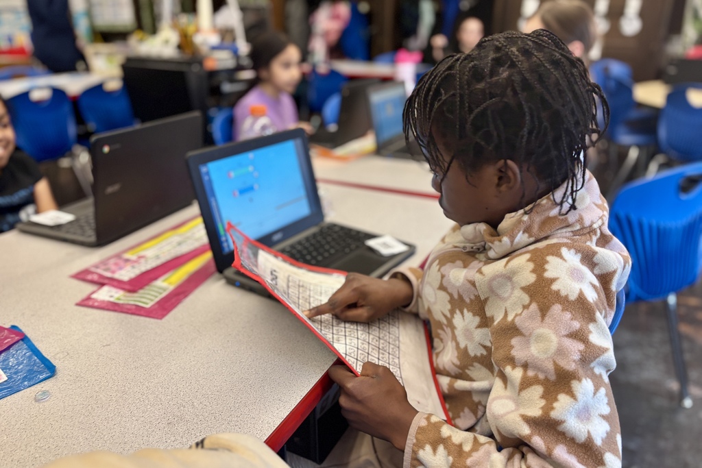 A young student with braided hair points to a worksheet while using a laptop in a classroom.