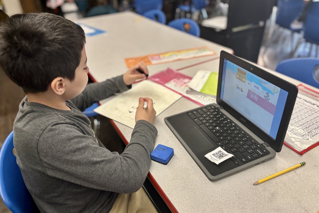 A young student uses a laptop and notebook for a math lesson in a classroom.