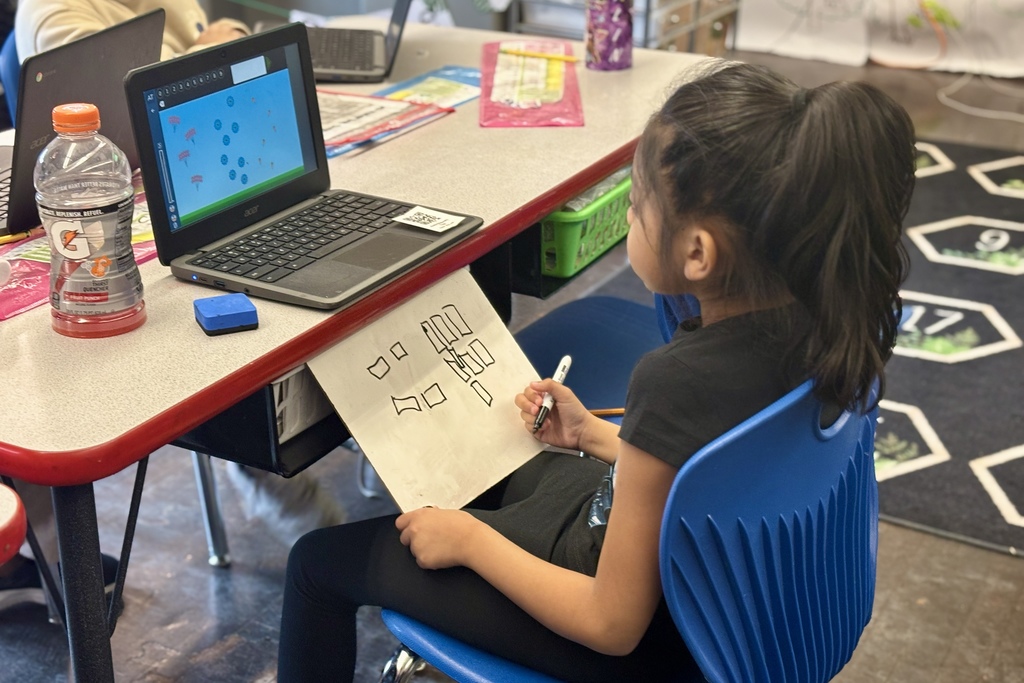 A young student uses a laptop and draws on a whiteboard at a classroom desk.