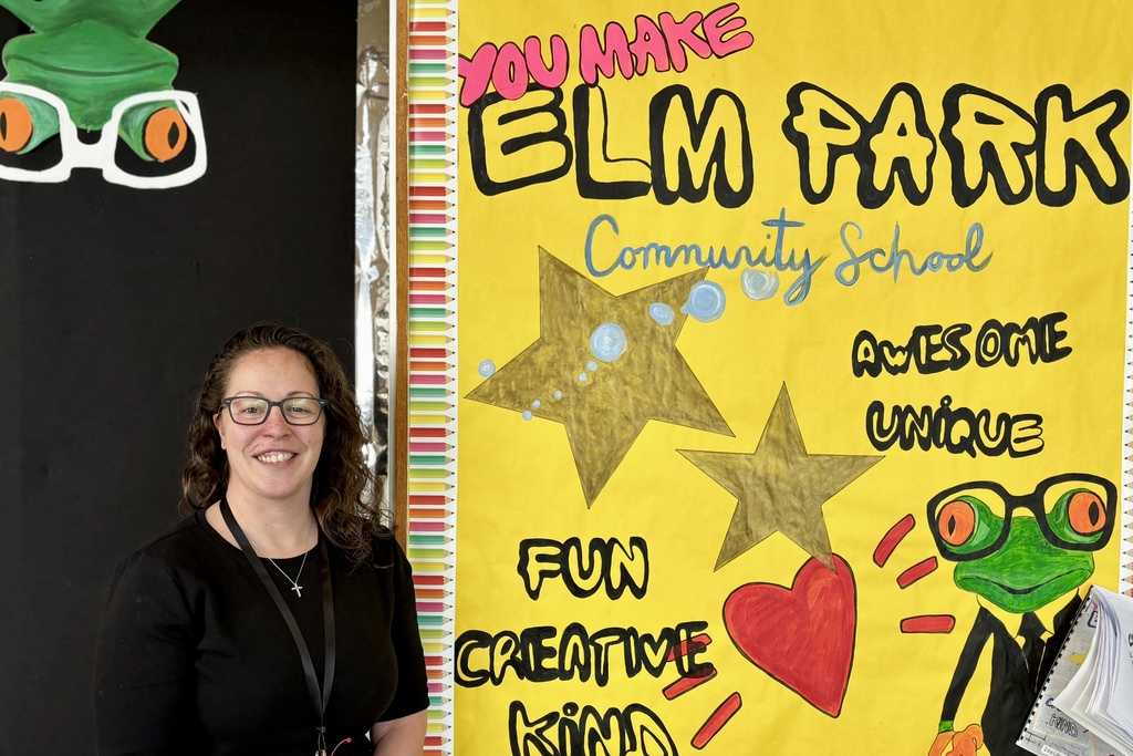 A smiling woman stands next to a colorful bulletin board that reads 'YOU MAKE ELM PARK Community School' with drawings of stars, a heart, and a frog.