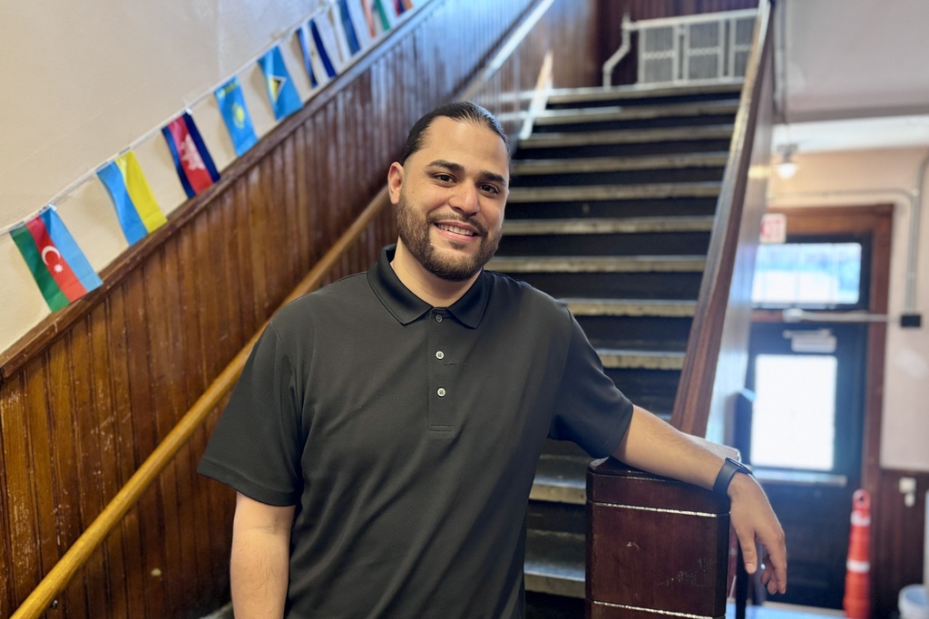 A smiling man in a black polo shirt leans on a wooden banister next to a staircase decorated with international flags.
