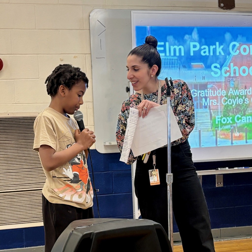 A young boy speaks into a microphone while a woman holds papers and smiles.