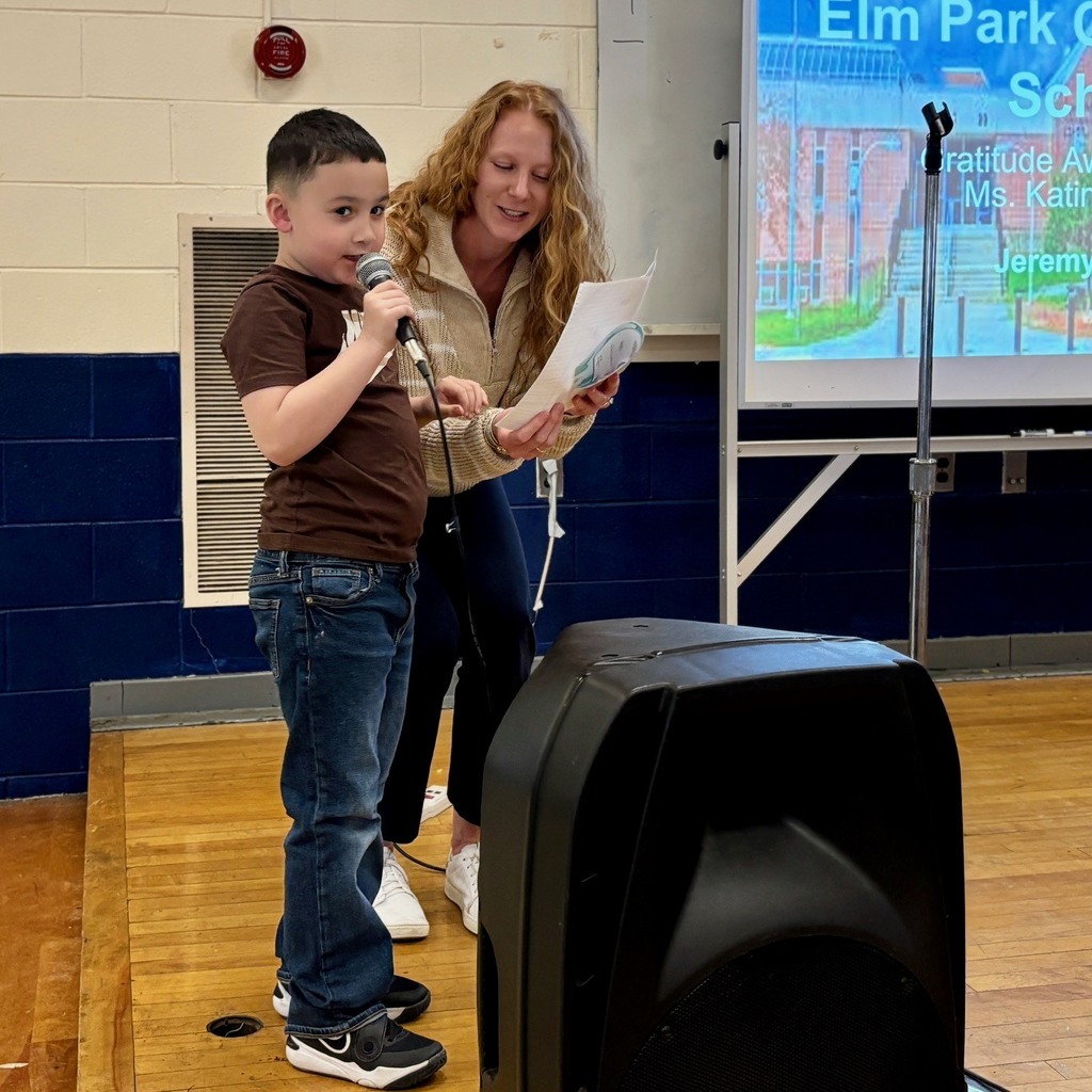 A young boy speaks into a microphone on a stage, with a woman holding papers beside him.