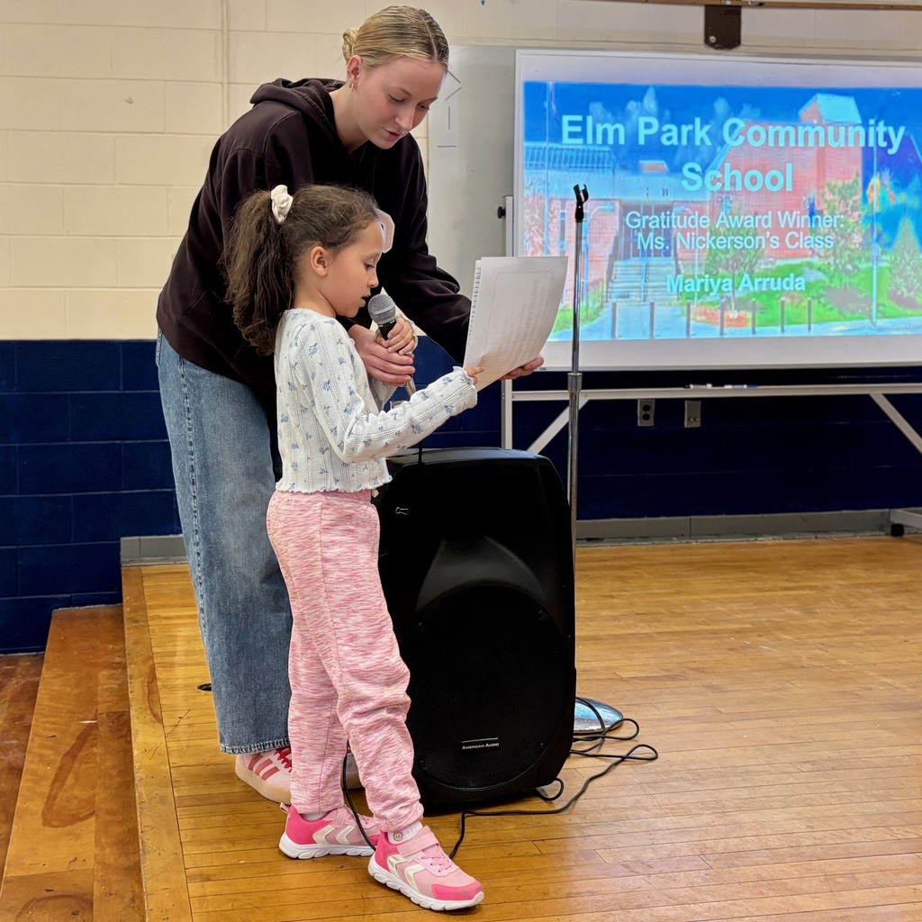 A young girl speaks into a microphone, with an adult assisting her, in front of a screen displaying "Elm Park Community School".