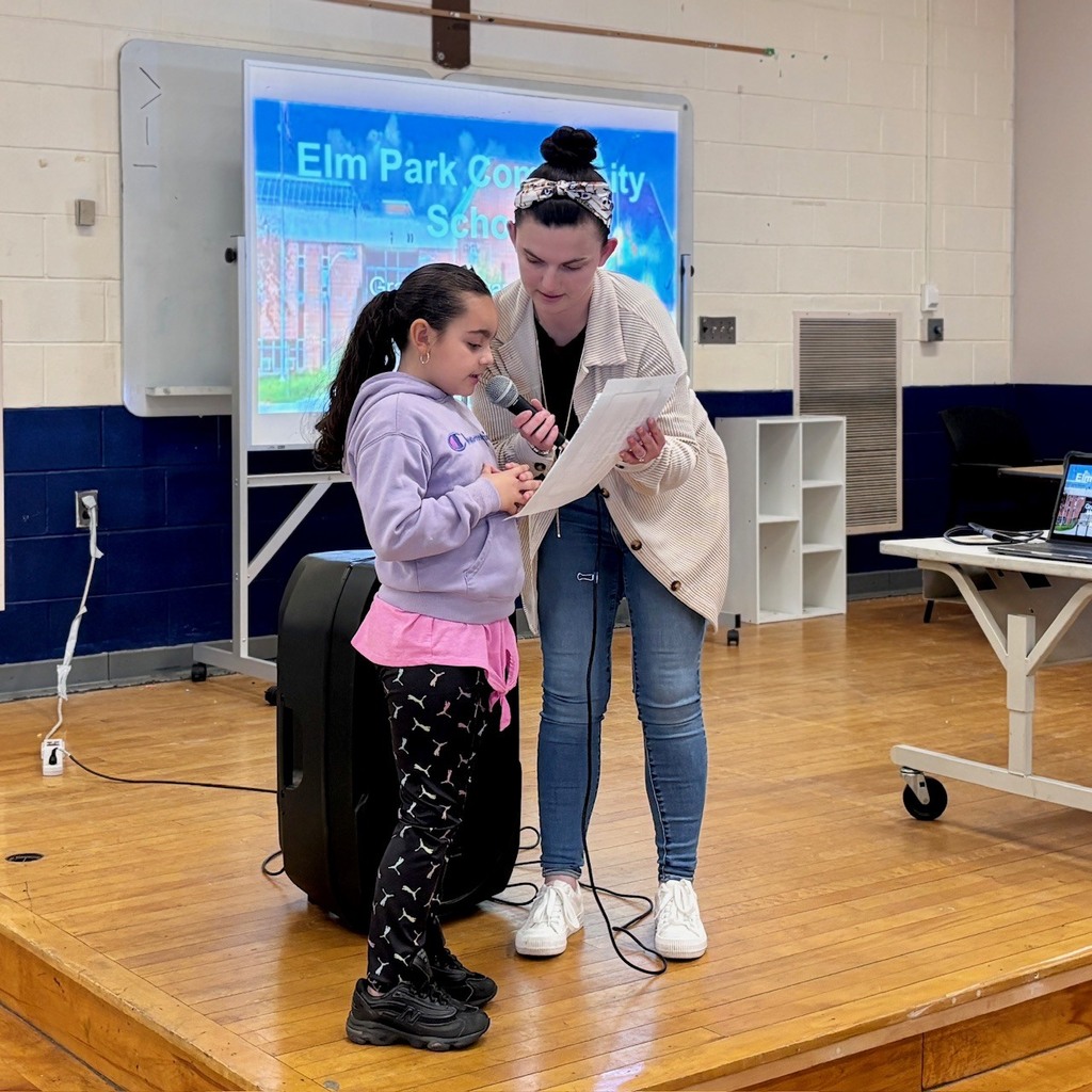 Two girls stand on a wooden stage, one holding a microphone and reading from a paper, the other assisting.