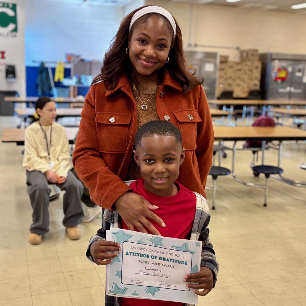 A smiling young boy holds an "Attitude of Gratitude" award from Elm Park Community School, with a woman standing behind him.