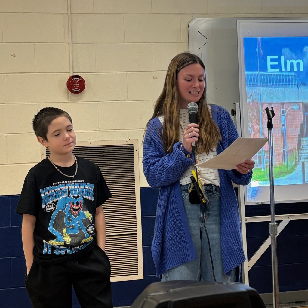 A young woman speaks into a microphone while holding a paper, standing next to a young boy.