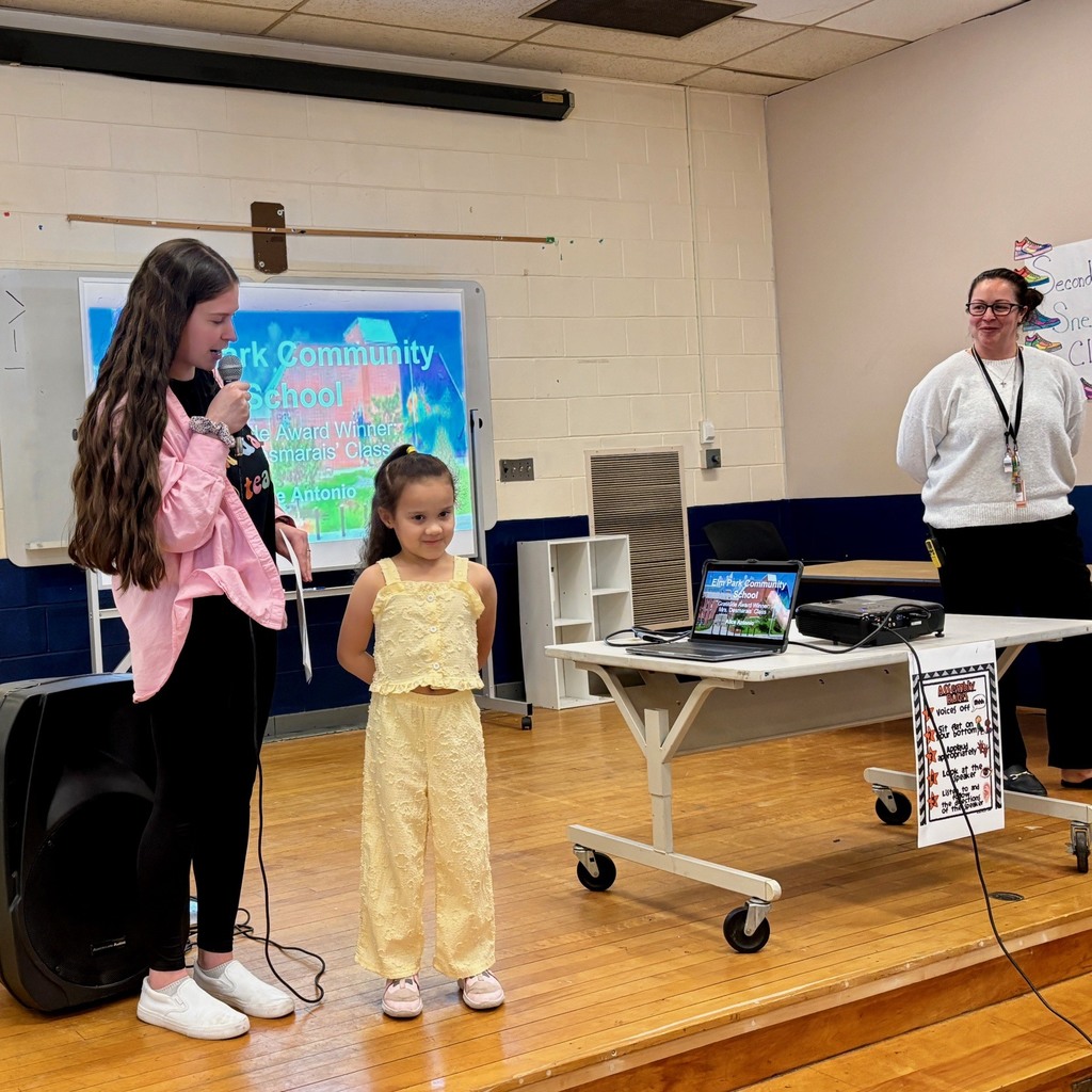 A young girl in a yellow outfit stands on a stage while a teen speaks into a microphone.