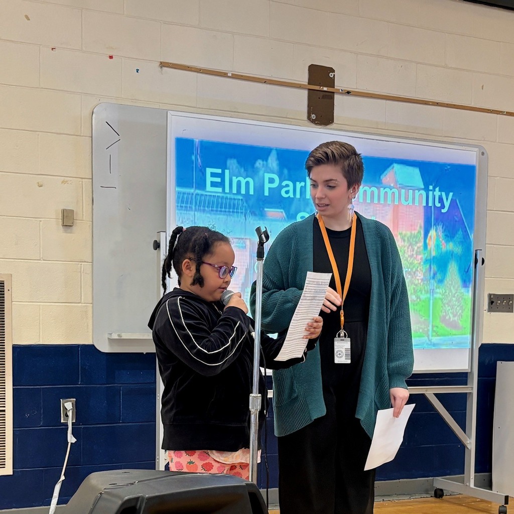 A young girl speaks into a microphone while an adult holds papers nearby, with "Elm Park Community" displayed on a screen.