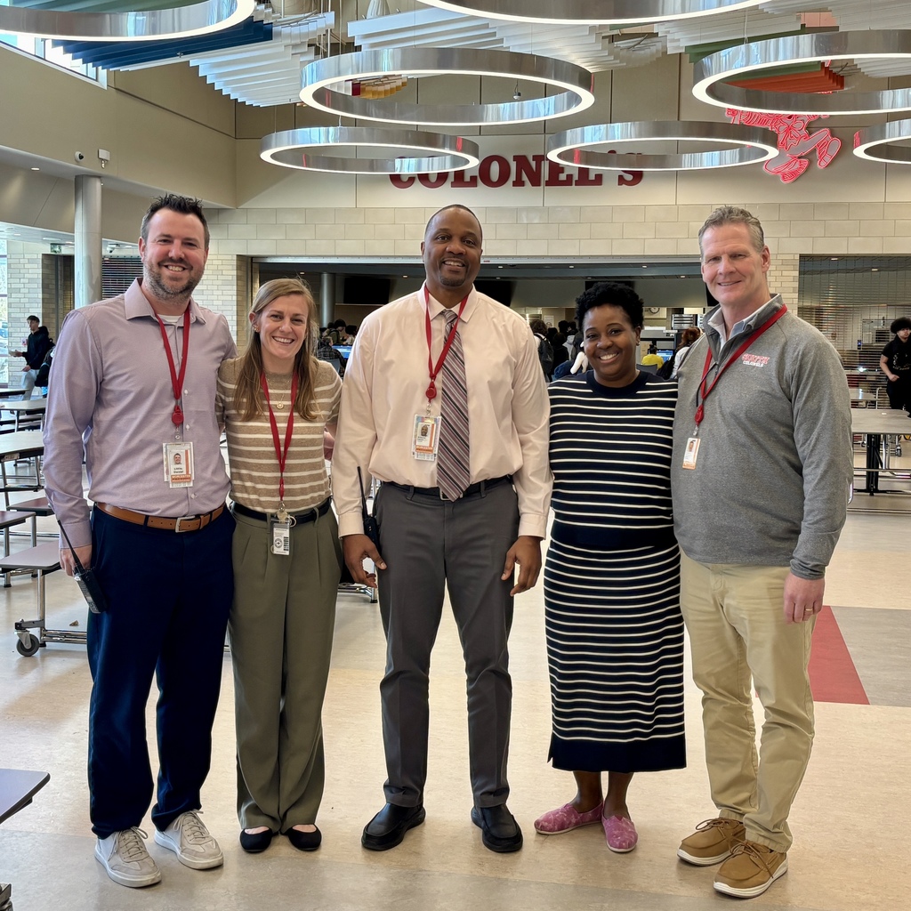 Five adults stand together in a school cafeteria, smiling at the camera.