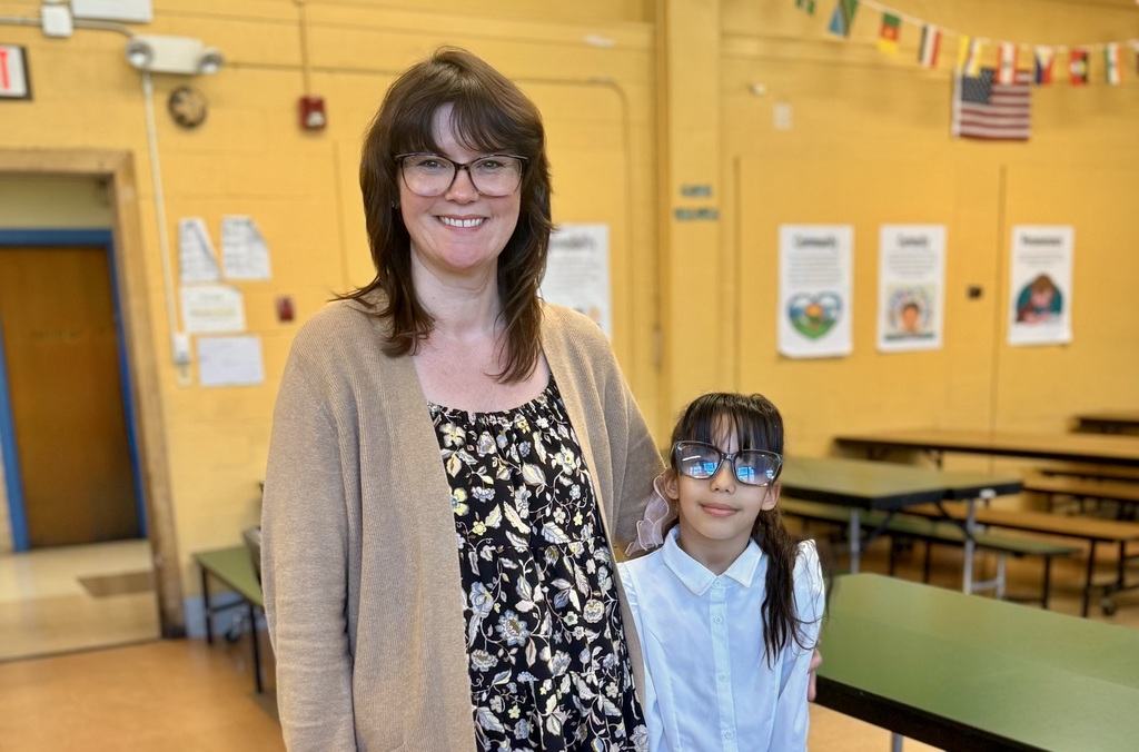A woman and a young girl stand together in a room with yellow walls, possibly a school cafeteria.