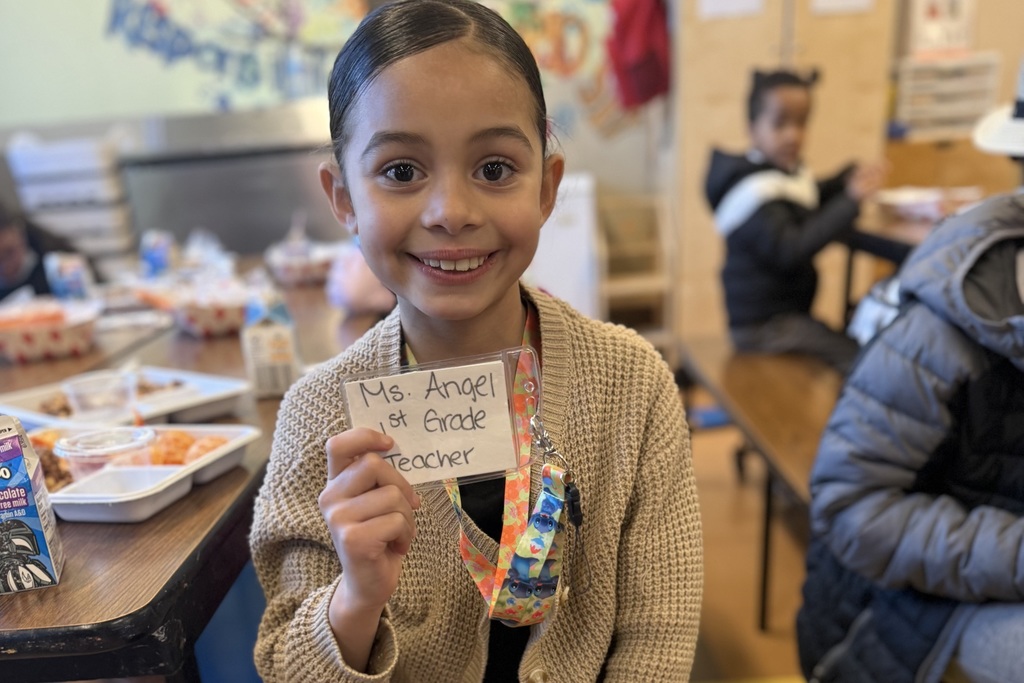 A young girl smiles while holding a name tag that reads 'Ms. Angel, 1st Grade Teacher'.
