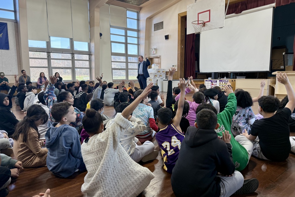 A man in a suit stands in front of a group of students sitting on the floor, many with their hands raised.