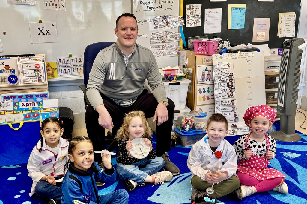 A teacher sits with five young children on a blue carpet in a classroom decorated with learning materials.