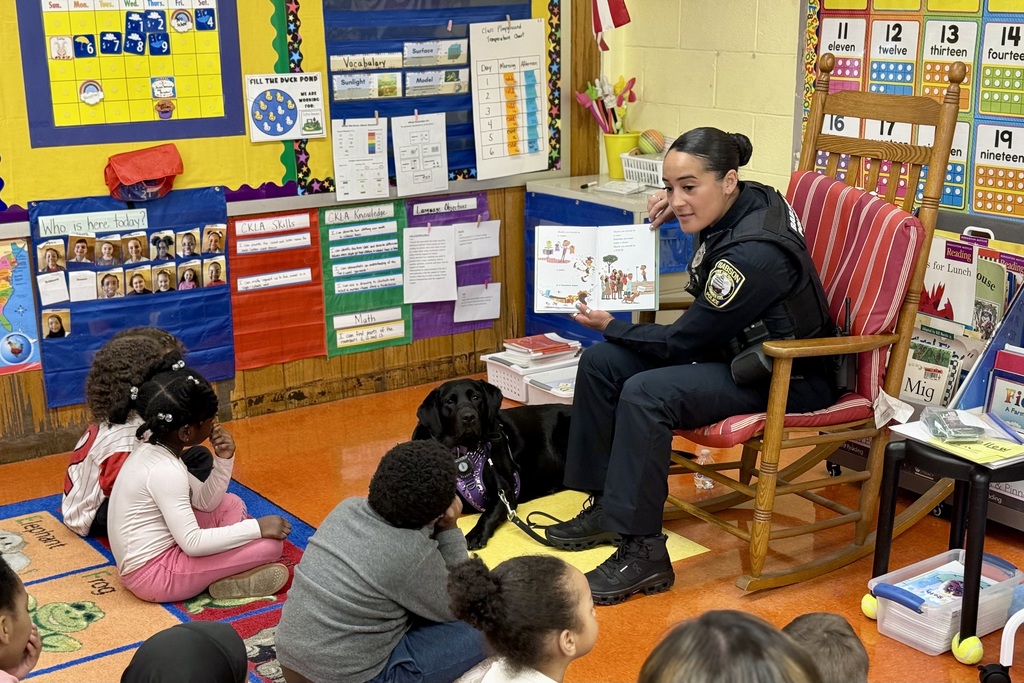 A police officer reads a book to a group of children and a black Labrador dog in a classroom.