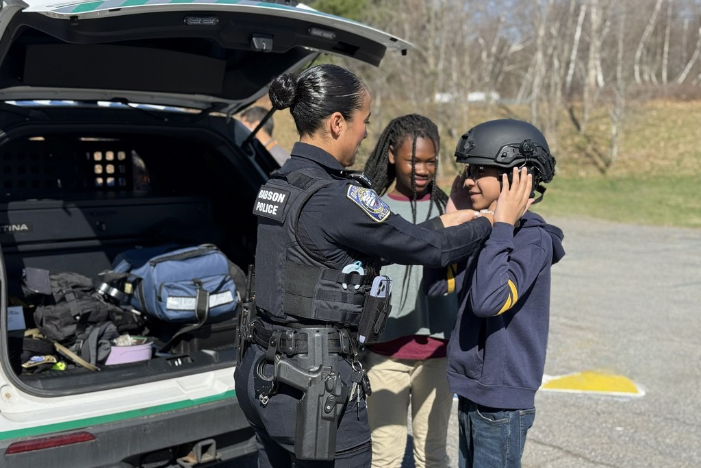 A police officer helps a child put on a tactical helmet near a police vehicle.