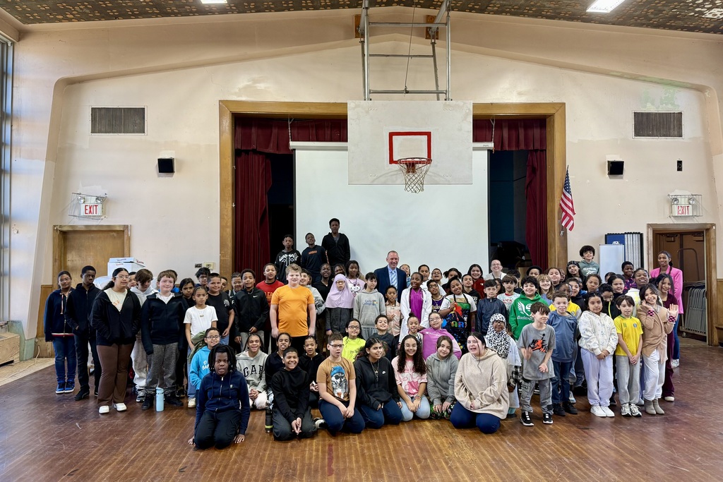 A large group of diverse students and adults pose for a photo in a gymnasium with a basketball hoop.