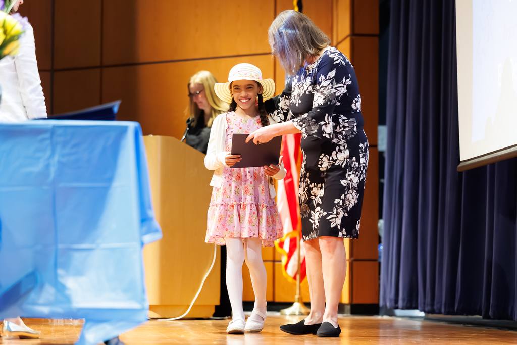 A student wearing a pink dress and a straw hat accepts a recognition certificate while smiling.