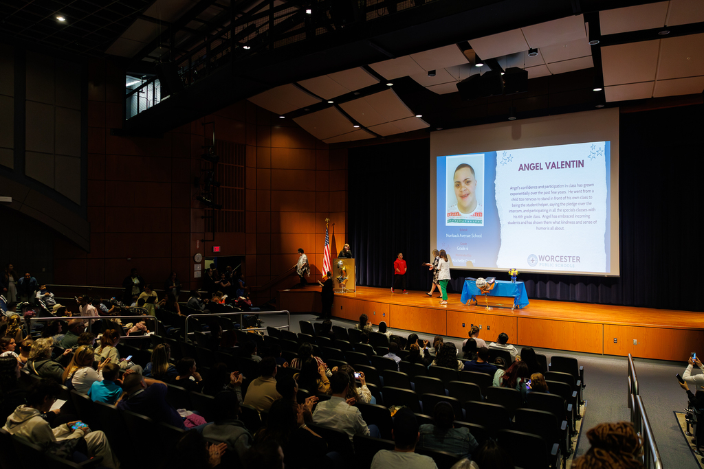 A student walks across a stage while accepting an recognition certificate, while patrons sit in the sits of the auditorium.