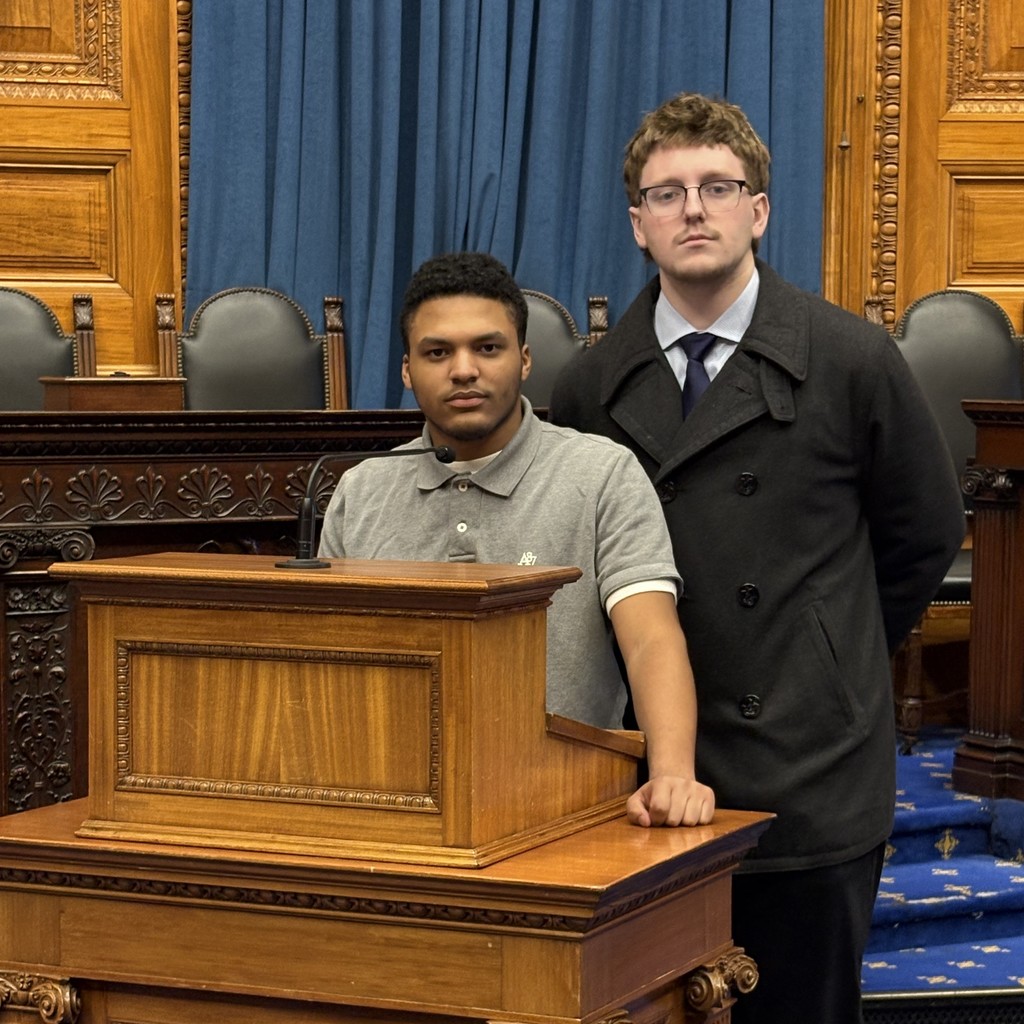 Two young men stand in a room with ornate wooden paneling and blue curtains, possibly a courtroom or legislative chamber.
