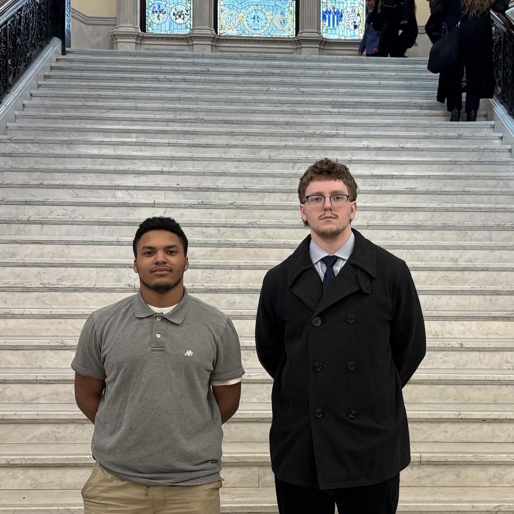 Two young men stand on a grand marble staircase with stained glass windows above.