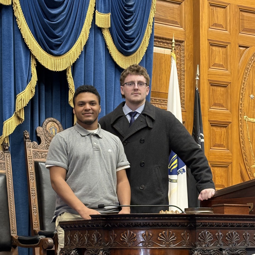 Two young men stand behind a wooden podium in a room with ornate blue curtains and wooden paneling.