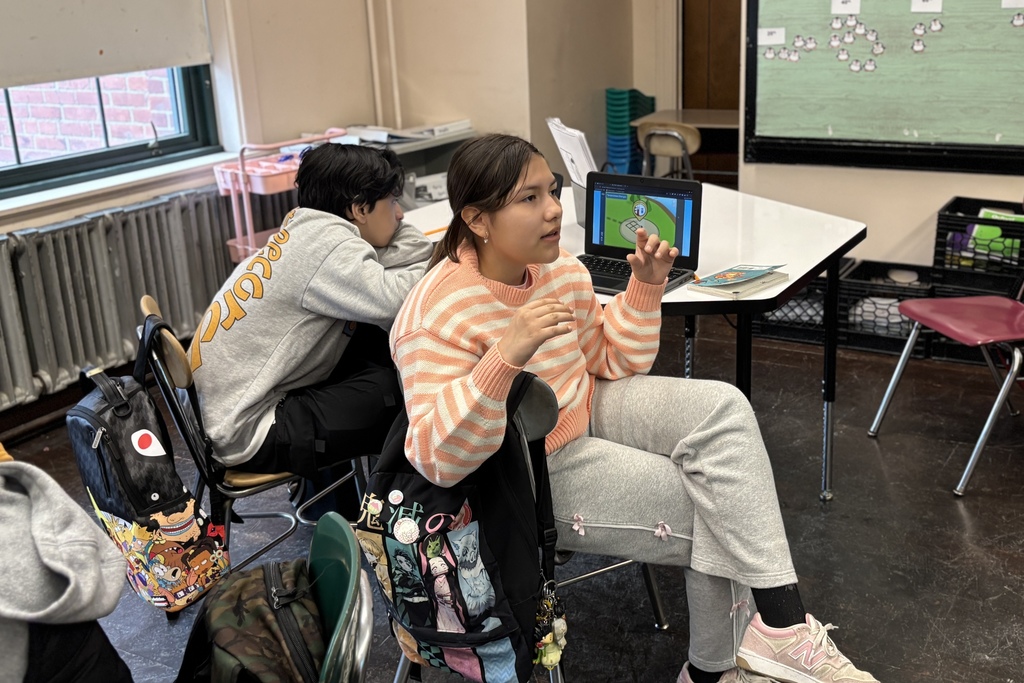 Two students are seated at desks in a classroom, with one looking at a laptop screen.