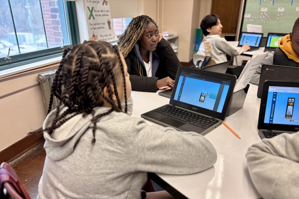 Students work on laptops in a classroom, with a whiteboard and posters visible.