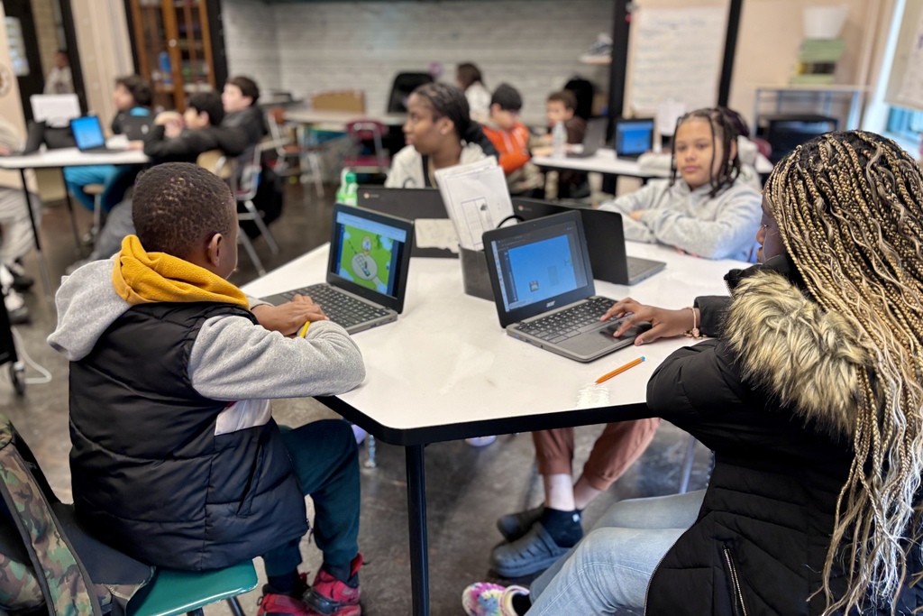 Students work on laptops at tables in a classroom setting.