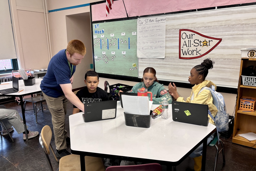 A teacher assists students working on laptops in a classroom with a "ST Math" board.