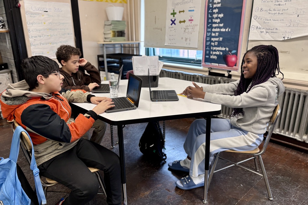 Three students work on laptops at a table in a classroom, with educational posters on the wall.