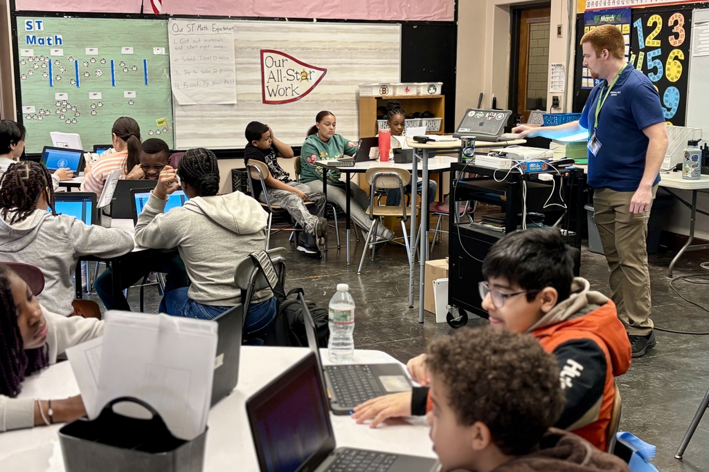 Students work on laptops in a classroom, with a teacher standing and observing.