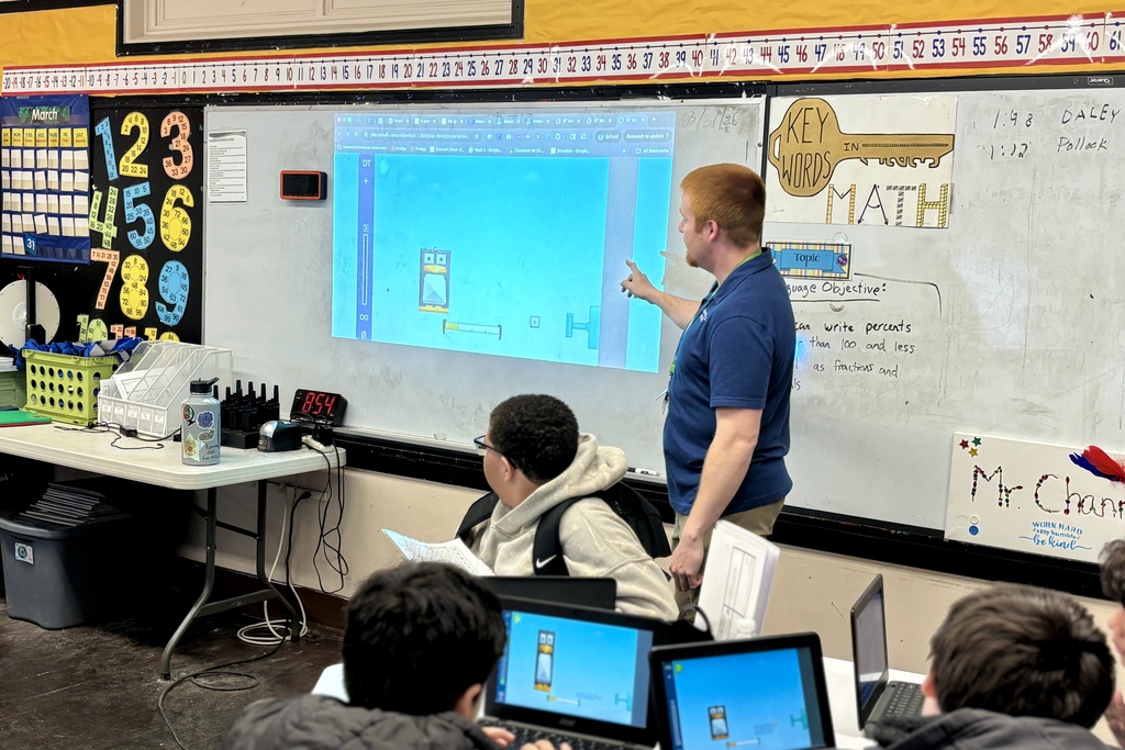 A teacher points to a projected game on a whiteboard in a classroom, with students looking on.