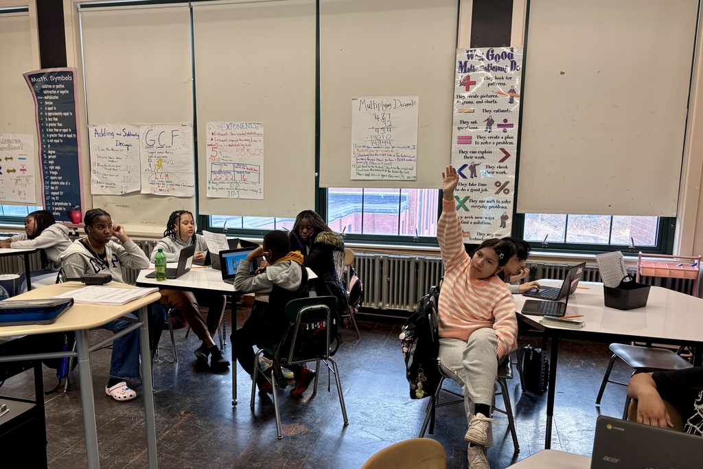 Students are seated at desks in a classroom, some looking at laptops and others at papers, with math-related posters on the wall.