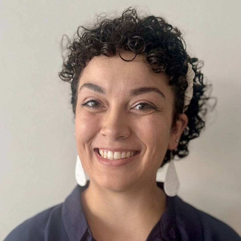 A woman with curly dark hair smiles warmly, wearing large white teardrop earrings and a dark blue shirt.