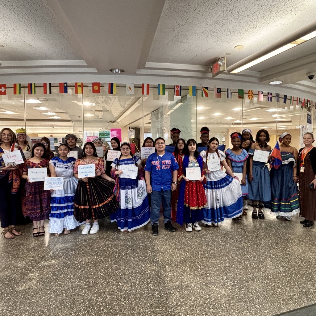A diverse group of people in traditional attire stand together, holding signs representing different countries.