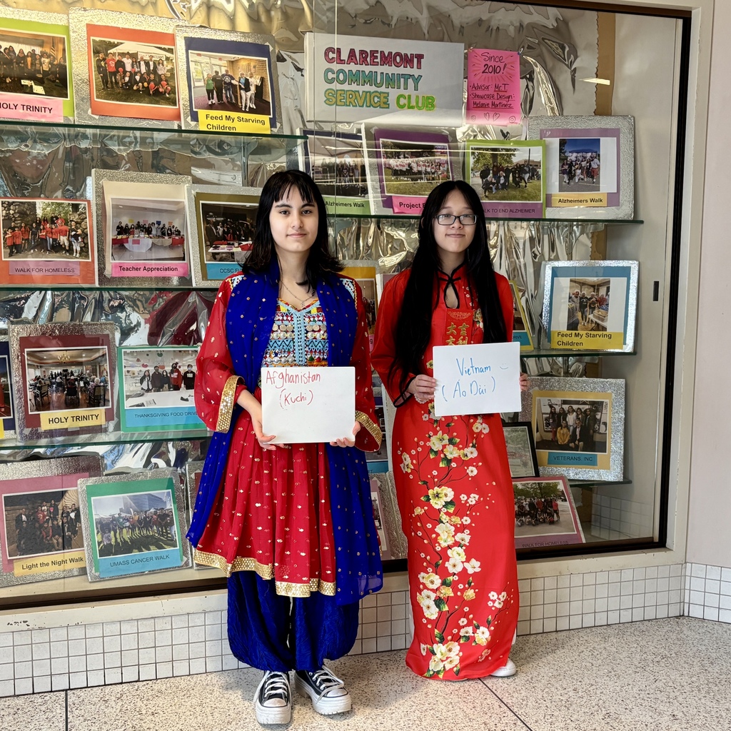 Two young women in traditional attire stand before a display of photos from the Claremont Community Service Club.