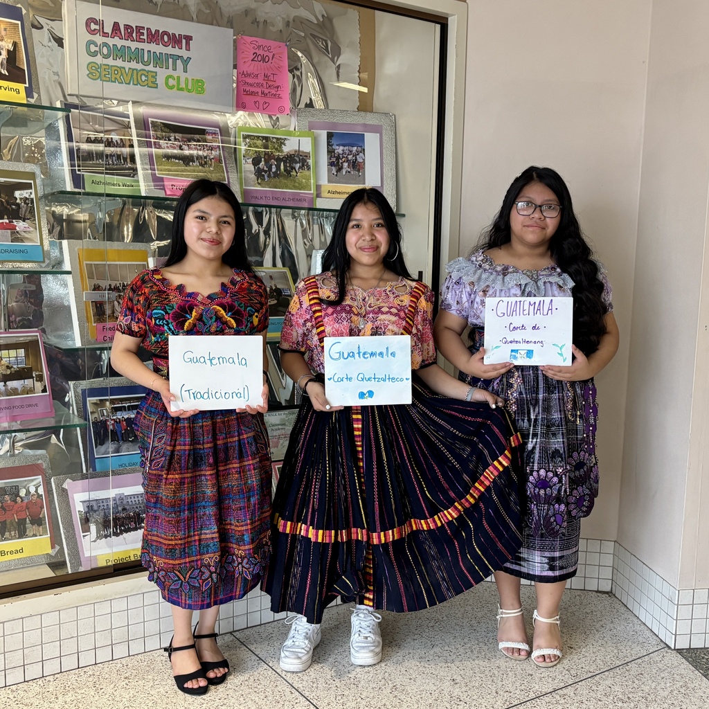 Three young women in traditional Guatemalan attire stand in front of a display board, each holding a sign identifying Guatemala.